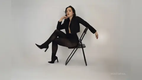 Elegant woman in a chic black blazer and tights sitting on a chair against a minimalist white background.
