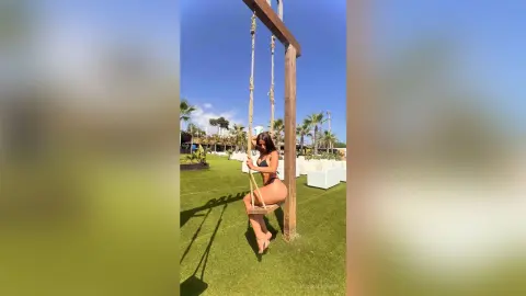 BiankaLeoness sits on a wooden swing outdoors in a black bikini, posing on the grass under a clear blue sky.