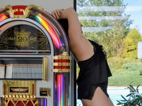 Woman in black lace loungewear leaning against a vintage jukebox near a sunny outdoor swimming pool.