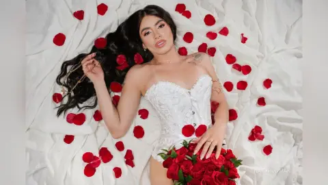 Woman in white lace corset lying on white sheets surrounded by red rose petals for a romantic mood.