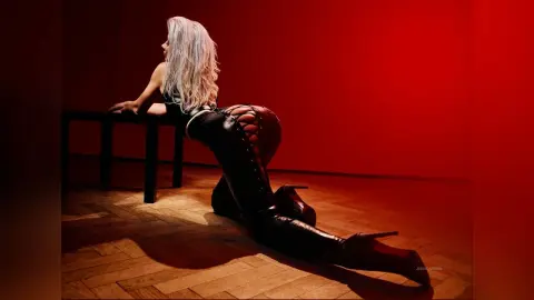 Elegant blonde woman in black lace-up leather pants posing on a wooden floor against a moody red background.