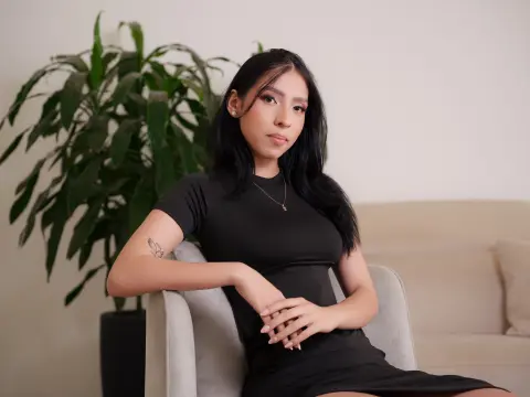 Young woman in a black dress sitting on a beige sofa next to a green houseplant in a bright, minimalist room.