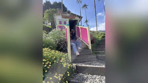 NatyGlow sits on wooden steps outside a colorful house, wearing a black tank top and light blue jeans, surrounded by white and yellow flowers.