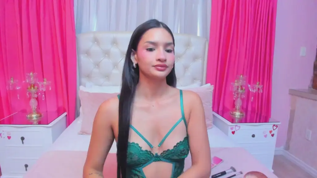 AuraClark wearing a green lace bra, posing in front of a white headboard and bright pink curtains.