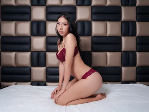 Young woman in burgundy lace lingerie kneeling on a white bed against a checkered headboard in a moody bedroom.