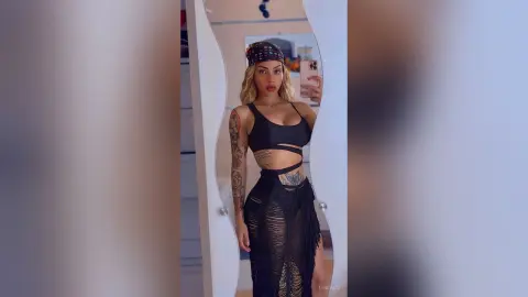 LindaDu poses in a black cut-out bikini top and a matching shredded skirt, showing off her blonde hair and arm tattoos in a mirror selfie.