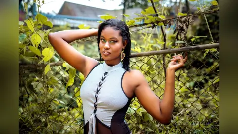 Smiling young woman in a grey sleeveless top posing against a lush green garden fence.