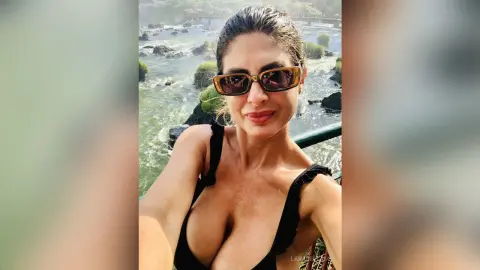 Smiling woman in black sunglasses and sleeveless top enjoying a sunny day by a rushing river waterfall.