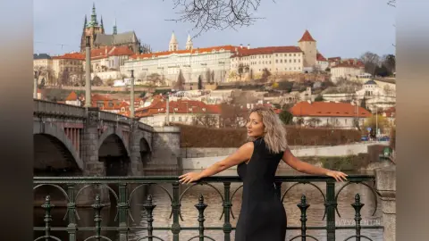 Woman in a black dress leaning on a bridge railing with Prague Castle in the background.