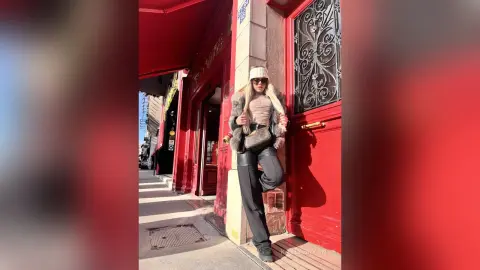 KaylaBryan poses in a white beanie, sunglasses, and a furry jacket while leaning against a red door on a sunny city sidewalk.