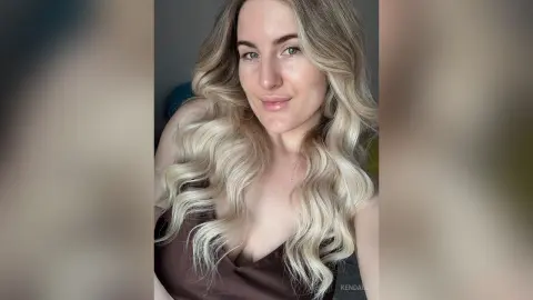 Blonde woman in a brown satin top smiling in a bright, minimalist indoor setting.
