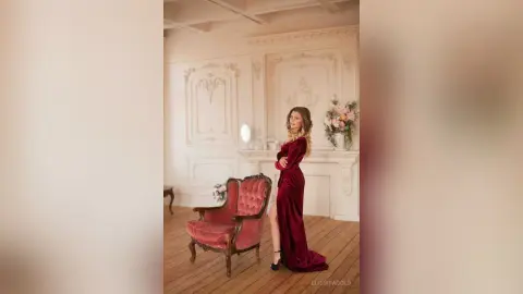 Elegant woman in a long red velvet gown posing in a classic vintage room with ornate white wall moldings.