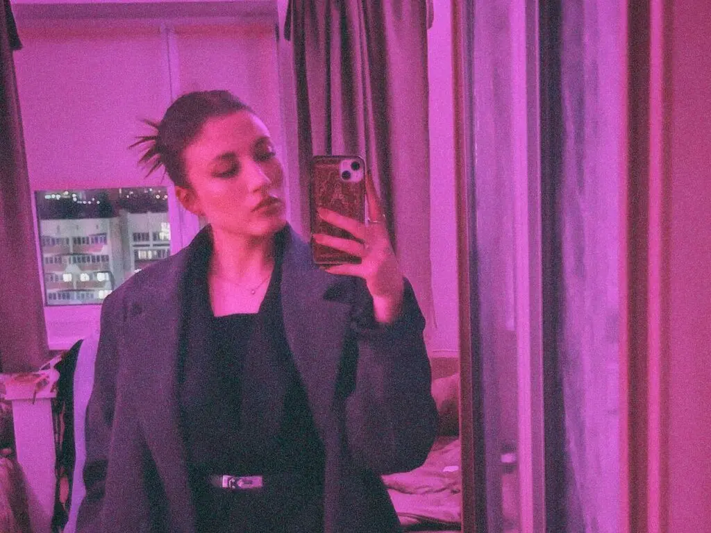 Young woman in a black outfit and blazer taking a moody mirror selfie in pink lighting.