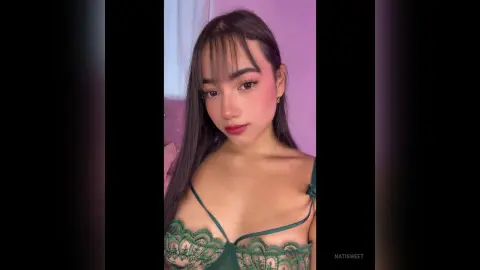 NatiSweet poses with long dark hair and heavy makeup, wearing a green lace bra or lingerie top.