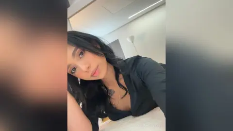 Young woman with long dark hair wearing a black top in a bright, modern bedroom setting.
