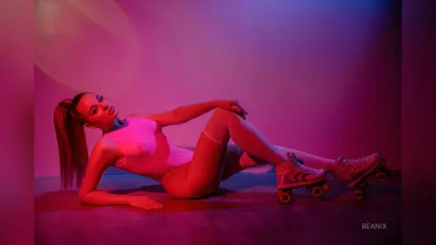 BeaNix reclines on the floor wearing a white bodysuit and roller skates under pink and blue studio lighting.