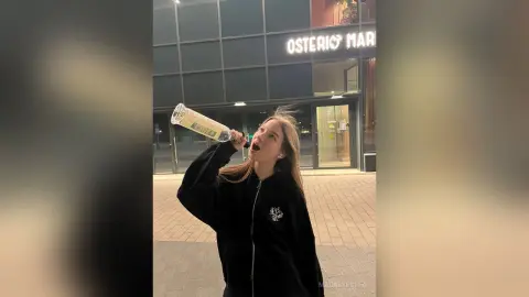 MagalyBoffa wearing a black zip-up hoodie, holding a glass bottle to her mouth while standing outside a building at night.