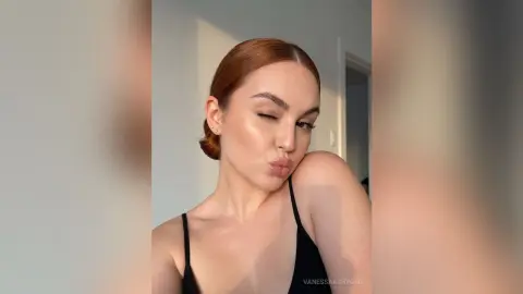 Redheaded woman in a black camisole making a playful kissy face in soft indoor lighting.