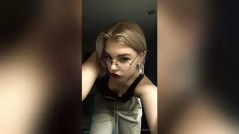 Blonde woman with glasses and dark lipstick posing in a black tank top for a moody indoor portrait.
