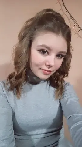 Young woman with wavy brown hair wearing a grey turtleneck sweater in a cozy indoor setting.