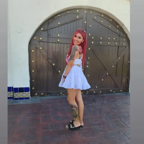 Smiling woman with red hair in a white summer dress standing before a rustic wooden arched door.