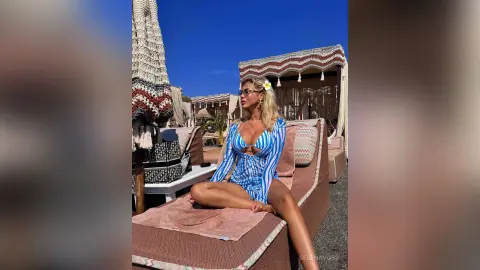 Blonde woman in a blue striped swimsuit relaxing on a sunbed at a luxury beach club under a sunny sky.
