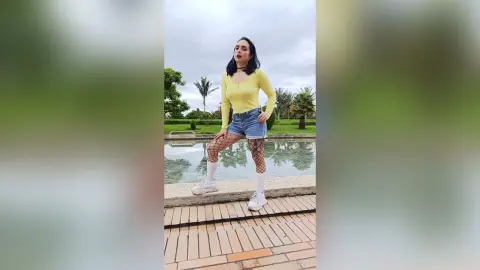 Confident woman in yellow top and denim shorts posing outdoors by a tranquil garden pond.