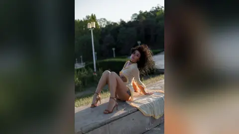 Elegant woman in a yellow summer dress sitting on a stone ledge in a lush green park during golden hour.