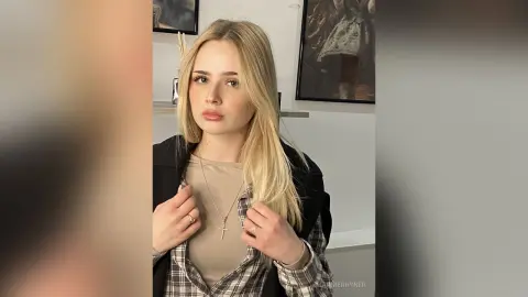 Blonde woman in a beige top and plaid shirt posing in a bright modern room.
