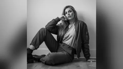 Hailey poses sitting on the floor wearing a dark bomber jacket, a light t-shirt, and dark pants with black boots.