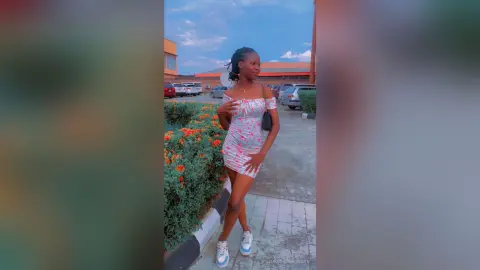 Young woman in a pink floral mini dress posing outdoors in a sunny parking lot setting.