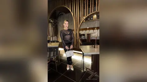 Elegant woman in a black sheer bodysuit and trousers posing in a modern, moody upscale restaurant.