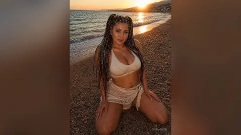 Brunette woman in white bikini sitting on a sandy beach during a golden sunset at the ocean.