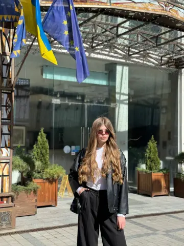 Stylish woman in a black leather jacket and white top standing outdoors near a modern building entrance.