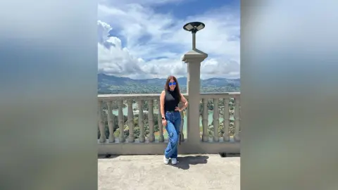 Smiling woman in a black tank top and blue jeans standing on a scenic balcony overlooking a mountain lake.