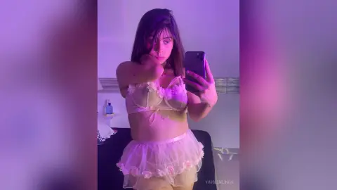 Young woman in white lace lingerie and a sheer ruffled mini skirt posing in soft purple mood lighting.