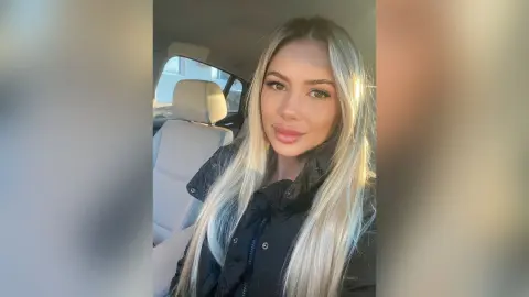 Blonde woman in a black winter jacket sitting in a car with warm sunlight lighting her face.