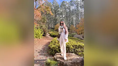 Stylish woman in white trousers and scarf walking through a sunny autumn forest path.