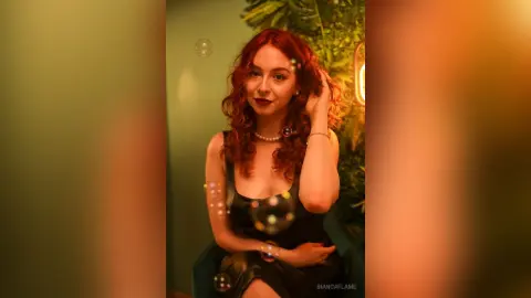 Redheaded woman in a black dress posing in a warm, moody indoor setting with floating bubbles.