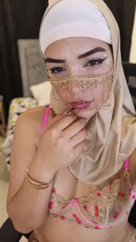 Woman in beige hijab and gold face jewelry wearing a pink lace bra in a bedroom setting.