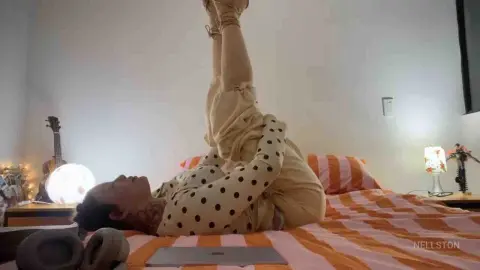 Woman in polka dot top doing yoga stretches on a bed in a cozy, brightly lit bedroom.