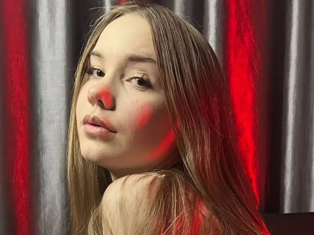 Young woman with long blonde hair posing against red and silver curtains in moody lighting.