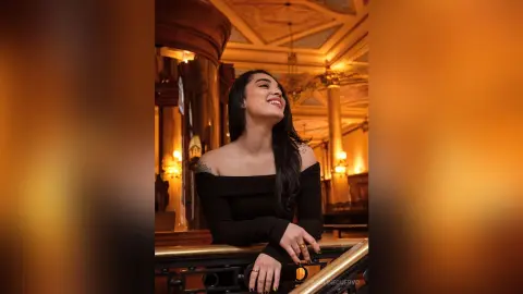 Smiling woman in a black off-the-shoulder top enjoying a warm, elegant indoor setting.