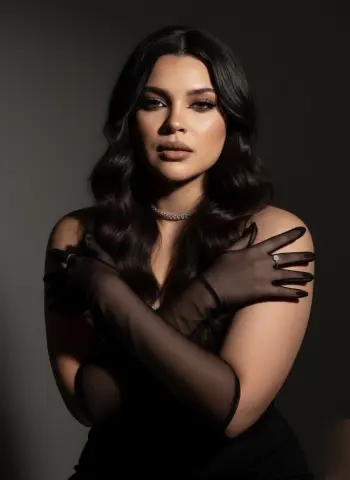 Elegant woman in a black dress and sheer gloves posing with a glamorous makeup look against a dark studio background.