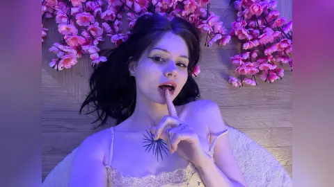 Young woman in white lace camisole lying on wood floor surrounded by pink cherry blossoms in moody purple lighting.