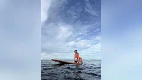 Young woman in a red swimsuit sitting on a surfboard in the ocean under a cloudy blue sky.