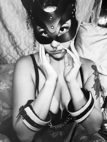 Woman in black leather mask and lingerie posing on a bed in a moody black and white portrait.