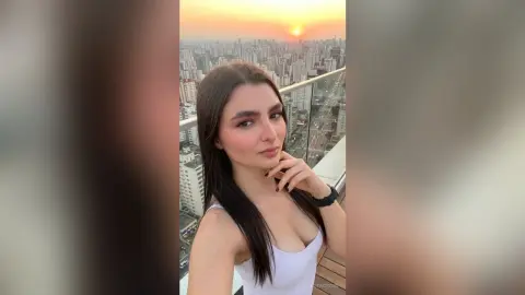 Young woman in a white tank top smiling on a city rooftop balcony during a golden sunset.