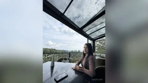 Young woman in a brown long-sleeve top sitting at a balcony table under a glass roof on a cloudy day.