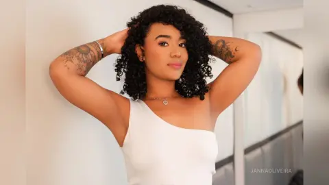 Smiling young woman with curly hair in a white one-shoulder top posing in a bright, modern indoor setting.